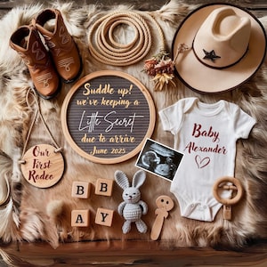 Könnte beinhalten: Ein Holzschild mit dem Text "Our First Rodeo" in roten Buchstaben, ein Schiefertafel-Schild mit dem Text "Suddle up! We've keeping a Little Secret due to arrive June 2025", ein weißer Strampler mit dem Text "Baby Alexander" in roten Buchstaben, eine Holzrassel, ein graues Häkelkaninchen und Holzklötze, die "BABY" buchstabieren.
