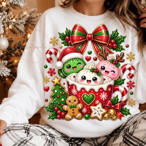Op de afbeelding: Een wit sweatshirt met een feestelijke afbeelding van drie lachende, ronde figuren in een rode mok, omringd door een peperkoekmannetje, een kerstboom, een grote rood-groene strik, zuurstokken en hulsttakken met rode bessen.