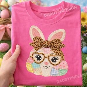Puede incluir: Camiseta rosa con un diseño de conejito con lazo de estampado de leopardo, gafas en forma de corazón y detalles de patchwork. El texto "DADDYPHDESIGN" está impreso en la camiseta.