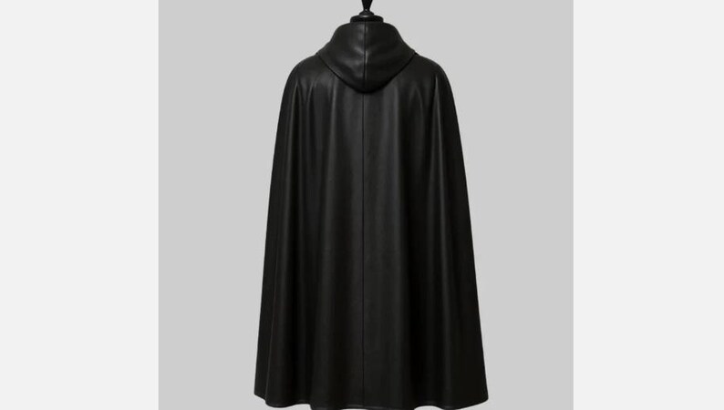 May include: A black hooded cape made of a smooth, possibly leather-like material. The cape is long, reaching to the ankles, and hangs from a black mannequin head. The back view shows the hood and the flowing fabric.