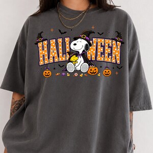 Puede incluir: Una camiseta gris carbón con un gráfico de Snoopy y Woodstock usando sombreros de bruja. El texto HALLOWEEN está impreso en letras grandes de color naranja con puntos blancos. Tres calabazas y dulces se muestran debajo.