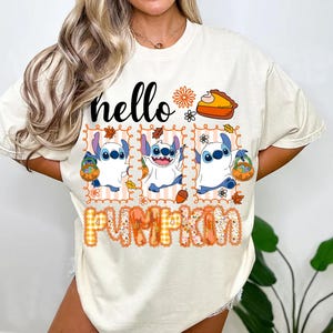 Puede incluir: Una camiseta de manga corta color crema con el texto hello PUMPKIN. El gráfico incluye tres paneles del personaje Stitch disfrazado de fantasma, sosteniendo un cubo de calabaza, rodeado de hojas de otoño, flores y un trozo de tarta.