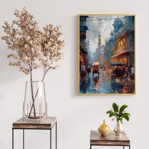 May include: A framed art print depicts a vibrant street scene with horse-drawn carriages and buildings with domes. The artwork features a palette of blues, oranges, and yellows, creating a sense of a bustling city. The frame is gold.