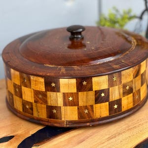 Op de afbeelding: Een ronde houten doos met een deksel, met een schaakbordpatroon met ingelegde messing sterren. De doos is gemaakt van donker hout met een natuurlijke afwerking.