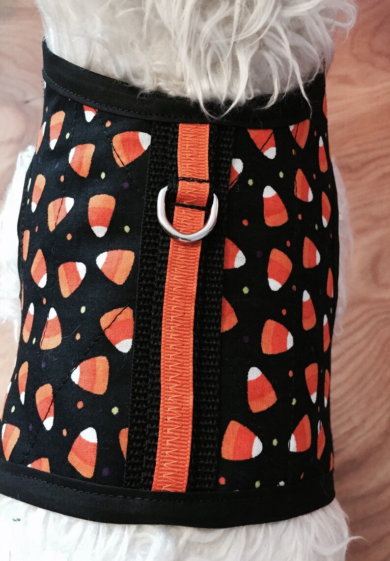 May include: A black dog harness with an orange and white candy corn pattern. The harness has a silver D-ring and an orange adjustable strap.