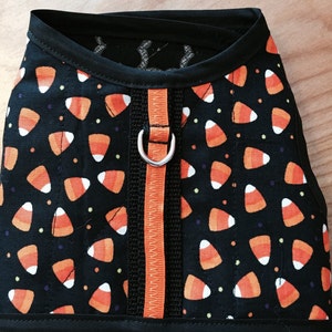 May include: A black dog harness with an orange and white candy corn pattern. The harness has a black strap with an orange zipper and a silver D-ring.