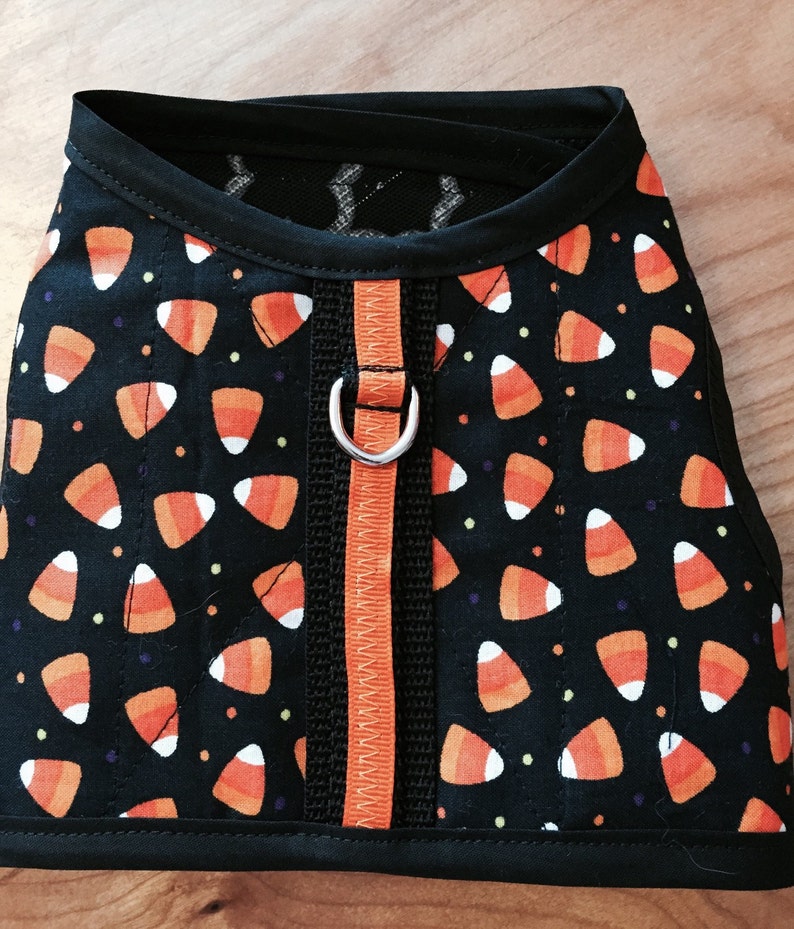 May include: A black dog harness with an orange and white candy corn pattern. The harness has a black strap with an orange zipper and a silver D-ring.