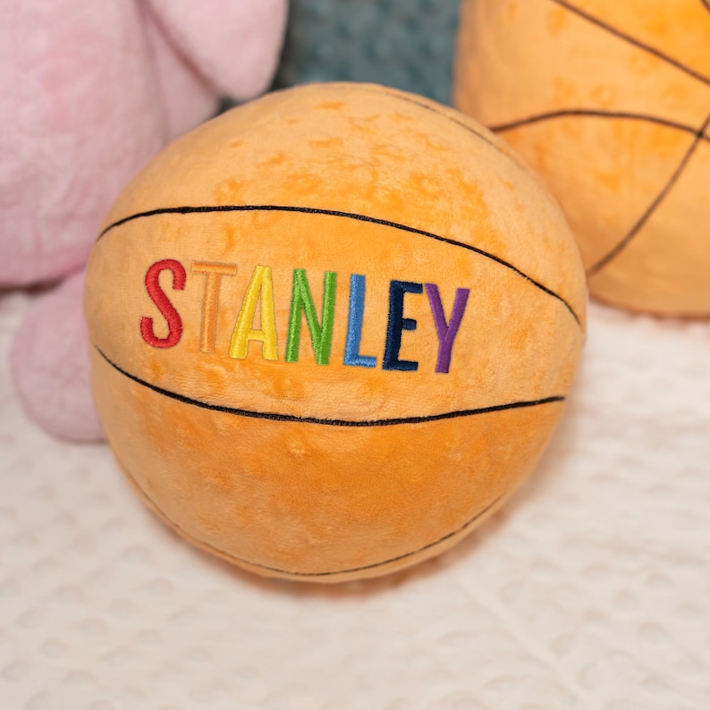 May include: A round, orange plush basketball with a textured surface and black stitched lines. The name STANLEY is embroidered across the center in multi-colored letters, with each letter featuring a different hue including red, yellow, green, blue, and purple.