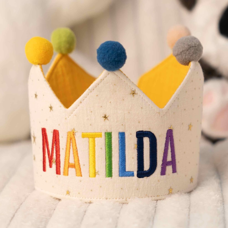 May include: A cream-colored fabric crown featuring a gold star pattern and the name MATILDA embroidered in rainbow-colored letters. The crown is topped with five colorful pom-poms in yellow, green, blue, peach, and gray, with a yellow lining.