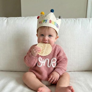 Może przedstawiać: Kremowa korona z materiału z kolorowymi pomponami i napisem HAPPY BIRTHDAY w kolorach tęczy, noszona do dzianinowego swetra w kolorze przybrudzonego r&oacute;żu z białym napisem one oraz okrągłą drewnianą tabliczką z napisem twelve months.