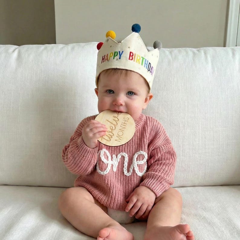 Może przedstawiać: Kremowa korona z materiału z kolorowymi pomponami i napisem HAPPY BIRTHDAY w kolorach tęczy, noszona do dzianinowego swetra w kolorze przybrudzonego r&oacute;żu z białym napisem one oraz okrągłą drewnianą tabliczką z napisem twelve months.