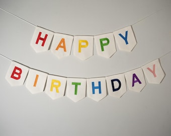 May include: A white banner with the words "HAPPY BIRTHDAY" in colorful embroidered letters. The letters are red, orange, yellow, green, blue, purple, and pink. The banner is strung on a thin white string.