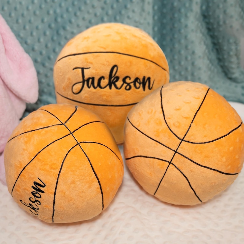 May include: Three round, orange, textured plush basketball pillows. Two of the pillows feature the name Jackson embroidered in black cursive script. The basketballs have black stitched lines detailing the classic ball pattern.