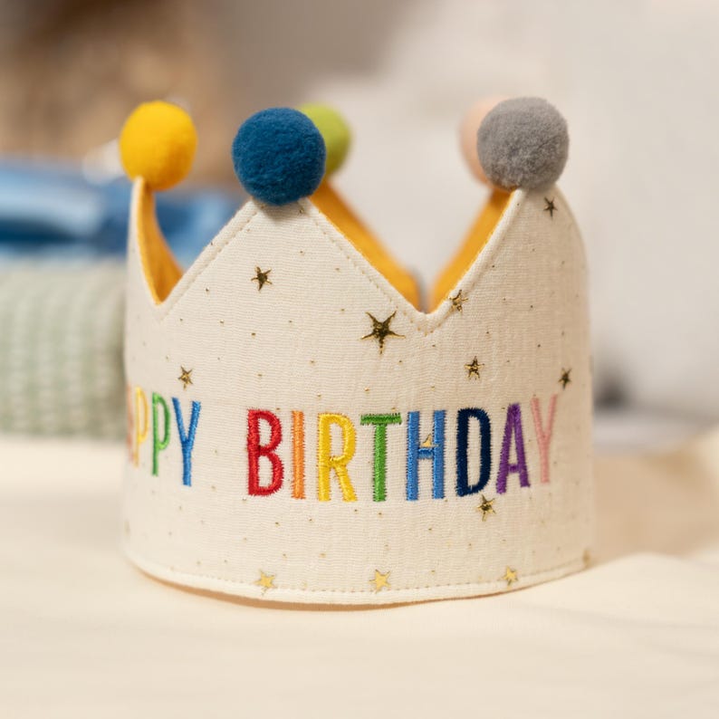 Może przedstawiać: Kremowa korona urodzinowa z tkaniny z wyhaftowanym napisem HAPPY BIRTHDAY wielokolorowymi literami. Korona jest ozdobiona małymi złotymi gwiazdkami i zwieńczona kilkoma puszystymi pomponami w kolorze ż&oacute;łtym, niebieskim, zielonym i szarym.