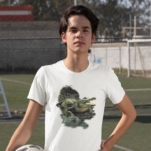 May include: A white t-shirt with a graphic print of a green alligator in a swampy setting. The person wearing the shirt is holding a soccer ball.