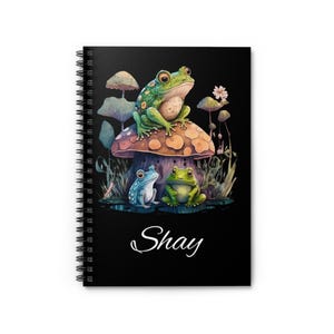 Peut inclure: Cahier à spirales noir avec une illustration fantaisiste de trois grenouilles. Une grande grenouille verte est assise sur un champignon, avec deux petites grenouilles en dessous. Le nom "Shay" est écrit en cursive.