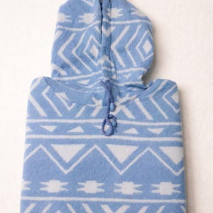 Peut inclure: Un sweat à capuche bleu clair plié avec un motif géométrique blanc. Le sweat à capuche a un cordon de serrage et une capuche. Le tissu semble doux et confortable. Le motif est un motif répétitif de triangles, de losanges et de zigzags.