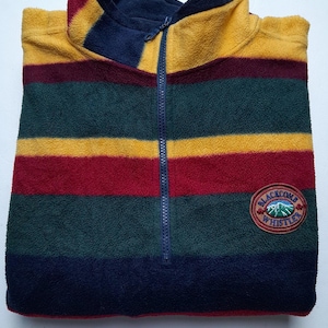 Könnte beinhalten: Ein gestreifter Fleece-Pullover in Gelb, Rot, Grün und Marineblau. Er hat einen marineblauen Kragen und einen Viertelreißverschluss. Ein runder Aufnäher mit Text und einer Berglandschaft befindet sich unten rechts. Bequemes Kleidungsstück.