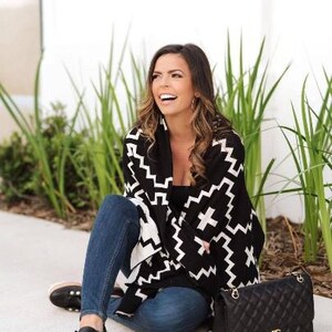 May include: A woman wearing a black and white patterned poncho with a black handbag. She is sitting on the ground with her legs crossed and smiling.