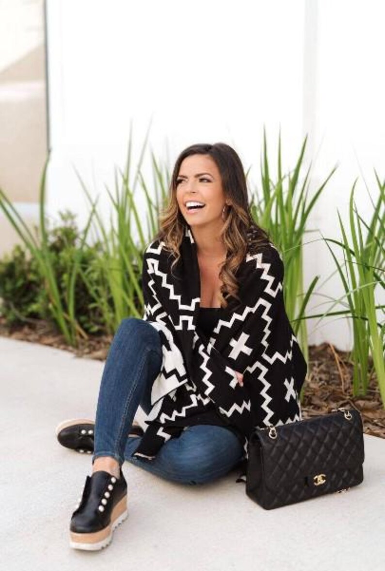 May include: A woman wearing a black and white patterned poncho with a black handbag. She is sitting on the ground with her legs crossed and smiling.