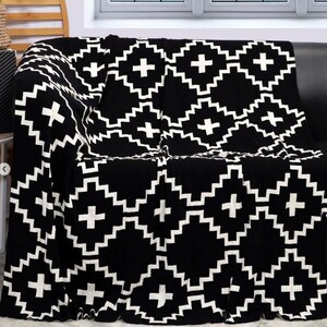 May include: Black and white throw blanket with a geometric pattern of plus signs and zigzags. The blanket is folded and draped over a black sofa.