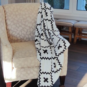 May include: A white armchair with a black and white geometric patterned throw blanket draped over it. The chair is in a room with a window and hardwood floors.