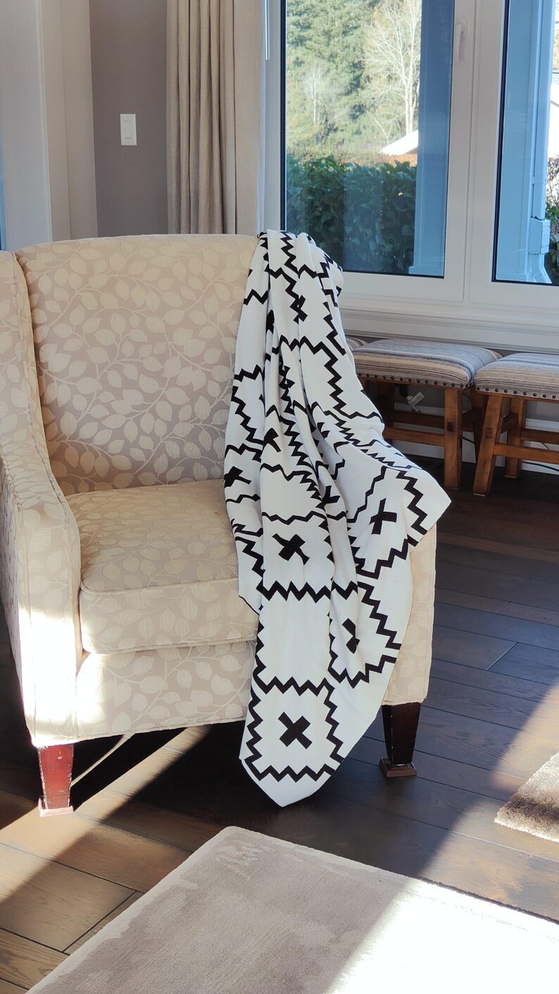 May include: A white armchair with a black and white geometric patterned throw blanket draped over it. The chair is in a room with a window and hardwood floors.