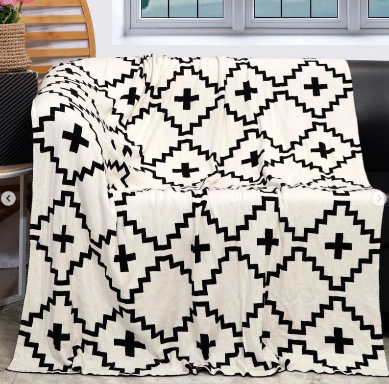 May include: A white throw blanket with a black geometric pattern of squares and crosses. The blanket is folded and draped over a couch.