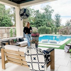 May include: A white and black patterned throw blanket draped over a wooden outdoor couch with white cushions. The couch is on a patio with a swimming pool in the background.
