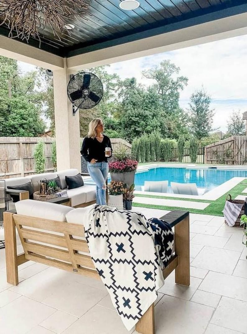 May include: A white and black patterned throw blanket draped over a wooden outdoor couch with white cushions. The couch is on a patio with a swimming pool in the background.