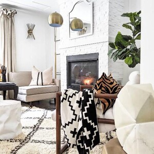May include: A white and black patterned throw blanket draped over a wooden chair with a white cushion and a tiger print throw pillow. The chair is in front of a fireplace with a black and white patterned rug on the floor.