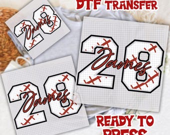 May include: Three DTF transfer sheets with a baseball-themed design. Each sheet features the number "28" in a black outline, with red stitching and the name "James" in a script font. The text "DTF TRANSFER" and "READY TO PRESS" are also visible.