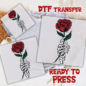 May include: DTF transfer sheets featuring a red rose held by a skeletal hand. The design includes green leaves and is printed on a grid-lined background. The text "DTF TRANSFER" and "READY TO PRESS" are also visible.