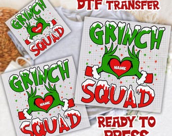 May include: DTF transfer sheets with a Grinch Squad design. The design features green text and Grinch hands forming a heart around a red heart with the word "NAME". The word "SQUAD" is in red with white snow accents.