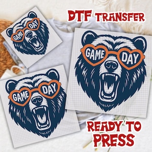Pu&ograve; includere: Design di trasferimento DTF con un orso blu navy che indossa occhiali da sole arancioni a forma di cuore con le parole "GAME DAY". L'orso ha la bocca aperta ed &egrave; pronto per essere pressato. Sono visibili anche le parole "DTF TRANSFER" e "READY TO PRESS".