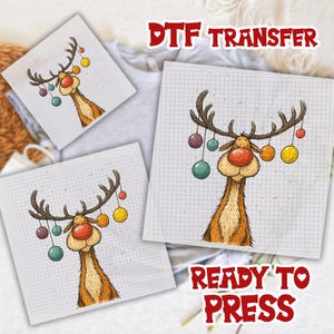 Op de afbeelding: DTF-transfervellen met een cartoon rendier met geweien versierd met kleurrijke ballen. Het rendier heeft een grote rode neus en staat tegen een rasterachtergrond. De tekst "READY TO PRESS" is ook zichtbaar.