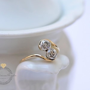 May include: A gold bypass ring with two round, clear gemstones. The ring's band curves around the gemstones, creating a unique and elegant design. The gemstones are set in a bezel setting, and the ring is displayed on a white surface.