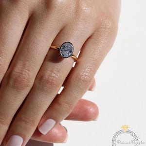 May include: A gold ring with an oval-shaped, light-colored gemstone set in a dark bezel. The ring is on a finger with a light pink nail polish. The ring is set against a white background.