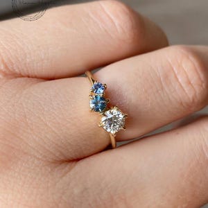 May include: A gold ring with three round stones. The center stone is a larger, white diamond. The two smaller stones on either side are blue sapphires.