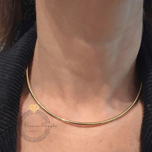 Può includere: Una collana girocollo color oro. La collana è una semplice, sottile fascia curva che si adatta vicino al collo. L'immagine mostra la collana su uno sfondo scuro.