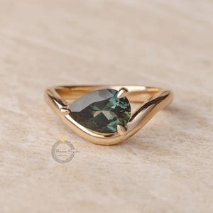 May include: A gold ring with a pear-shaped, teal-colored gemstone. The ring's band curves in a V-shape, with the gemstone held in place by two prongs. The ring is set against a neutral background.