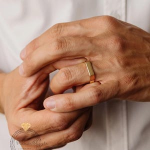May include: A gold rectangular ring is displayed on a finger. The ring has a flat top and sides, with a polished finish. The ring is shown against a white background.