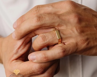 Könnte beinhalten: Ein goldfarbener, rechteckiger Ring wird an einem Finger getragen. Der Ring hat eine flache Oberseite und Seiten mit poliertem Finish. Der Ring wird vor einem weißen Hintergrund gezeigt.