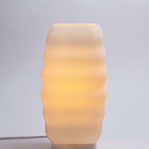 以下が含まれることがあります： クリーム色のリブ付きセラミックテーブルランプ。 ランプは、垂直の溝と内側から発せられる暖かく輝く光を備えた、波状の円筒形をしています。 ランプは白いコードで接続されています。
