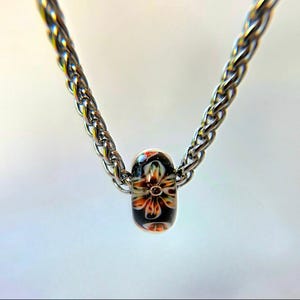 May include: A silver-toned chain necklace with a floral bead pendant. The bead features a black background with a colorful flower design in shades of orange, yellow, and white. The chain has a braided texture.