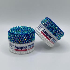 Puede incluir: Dos envases decorados de pomada curativa Aquaphor. Las tapas están cubiertas de pedrería azul iridiscente. Los envases son blancos con el nombre de la marca y la información del producto. Para pieles secas y agrietadas.