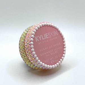 Op de afbeelding: Een roze Kylie Skin Sugar Lip Scrub-container met iriserende strass-steentjes en witte parelachtige kralen. De container heeft de tekst "KYLIESKIN BY KYLIE JENNER SUGAR LIP SCRUB GOMMAGE LEVRES AU SUCRE NET WT .35 OZ e10g."