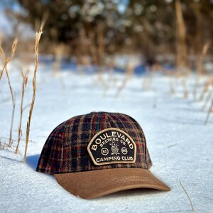 May include: A brown and black plaid baseball cap with a brown brim. The cap has a patch on the front that says "Boulevard Brewing Co. Camping Club" with a campfire graphic and the letters "KC" and "MO".
