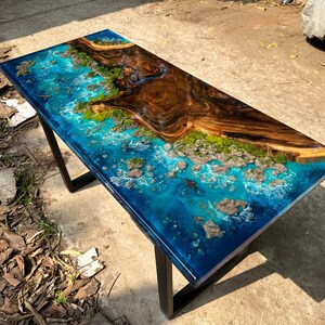 Puede incluir: Mesa rectangular de resina epoxi con una superficie azul oscuro, simulando una escena oce&aacute;nica con una isla de madera. La mesa tiene una base de metal negro y presenta una detallada representaci&oacute;n de olas y rocas.