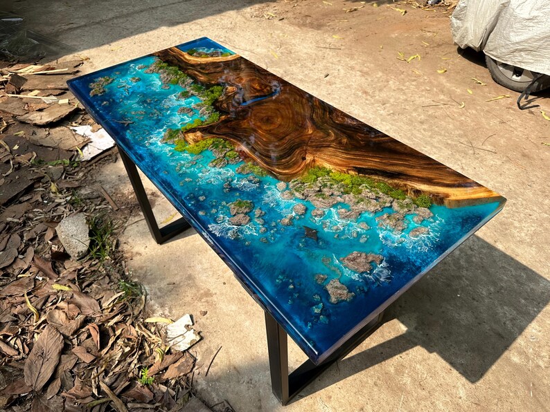 Puede incluir: Mesa rectangular de resina epoxi con una superficie azul oscuro, simulando una escena oce&aacute;nica con una isla de madera. La mesa tiene una base de metal negro y presenta una detallada representaci&oacute;n de olas y rocas.